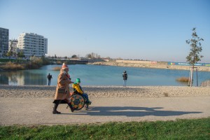 Frau mit Kind im Rollstuhl auf einem Gehweg. Im Hintergrund: der See. 